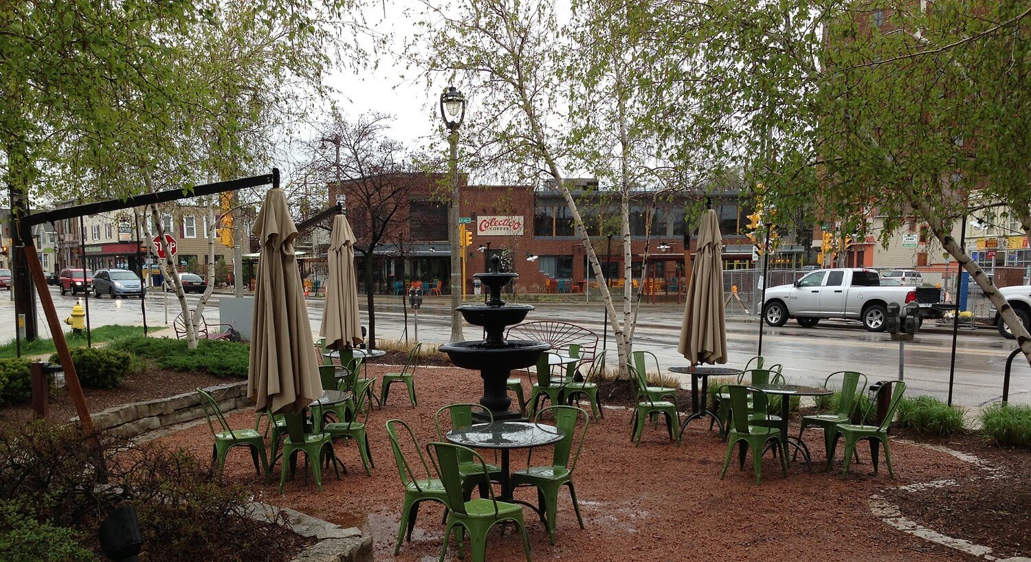 Seating area with green metal tables and chairs on a red gravel patio next to the street.
