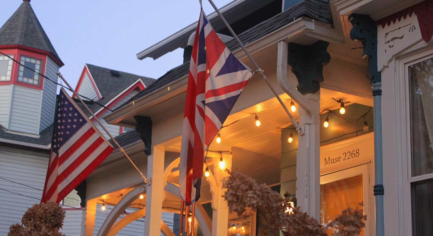 USA and UK flags fly from the porch at the main entrance. Warmly glowing white string lights decorate the porch ceiling. The turret and roof of the Victorian house next door are visible in the background. 