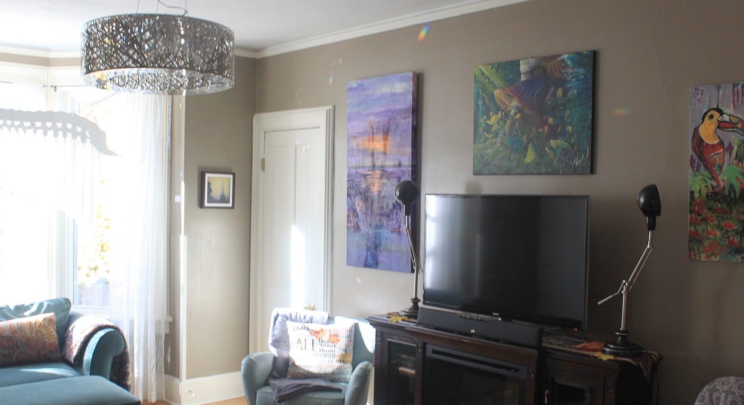 A dark beige wall filled with colorful paintings, and a TV on top of a fake fireplace. A decorative silver metal chandelier and a white door.