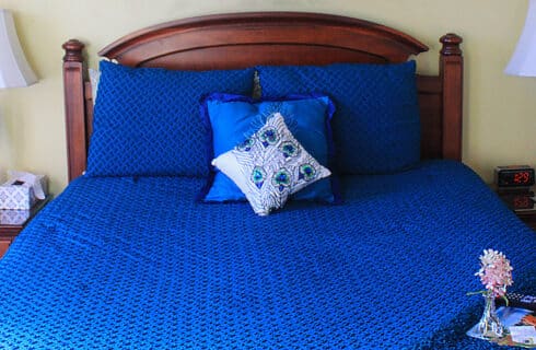 A cherrywood queen bed with peacock-blue bedspread and pillows in a room with yellow walls. Two night stands with lamps. On the end of the bed is a small tray with a flower vase, a key chain, and a TV remote control.