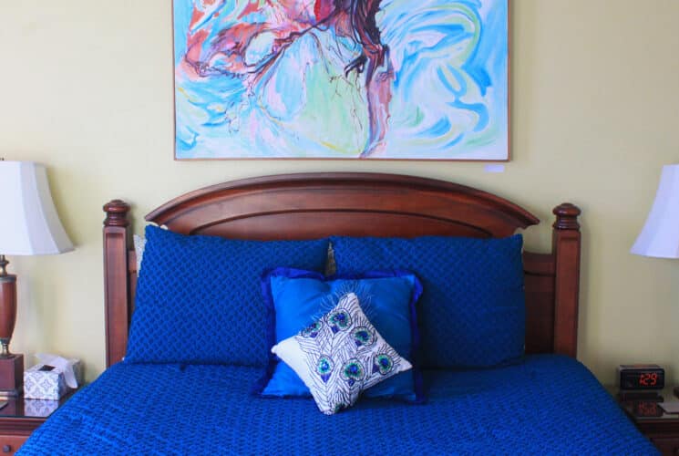 A queen bed with dark brown headboard and peacock blue bedspread in a room with pale yellow walls. A painting of a woman in a pool hangs on the wall above the headboard.