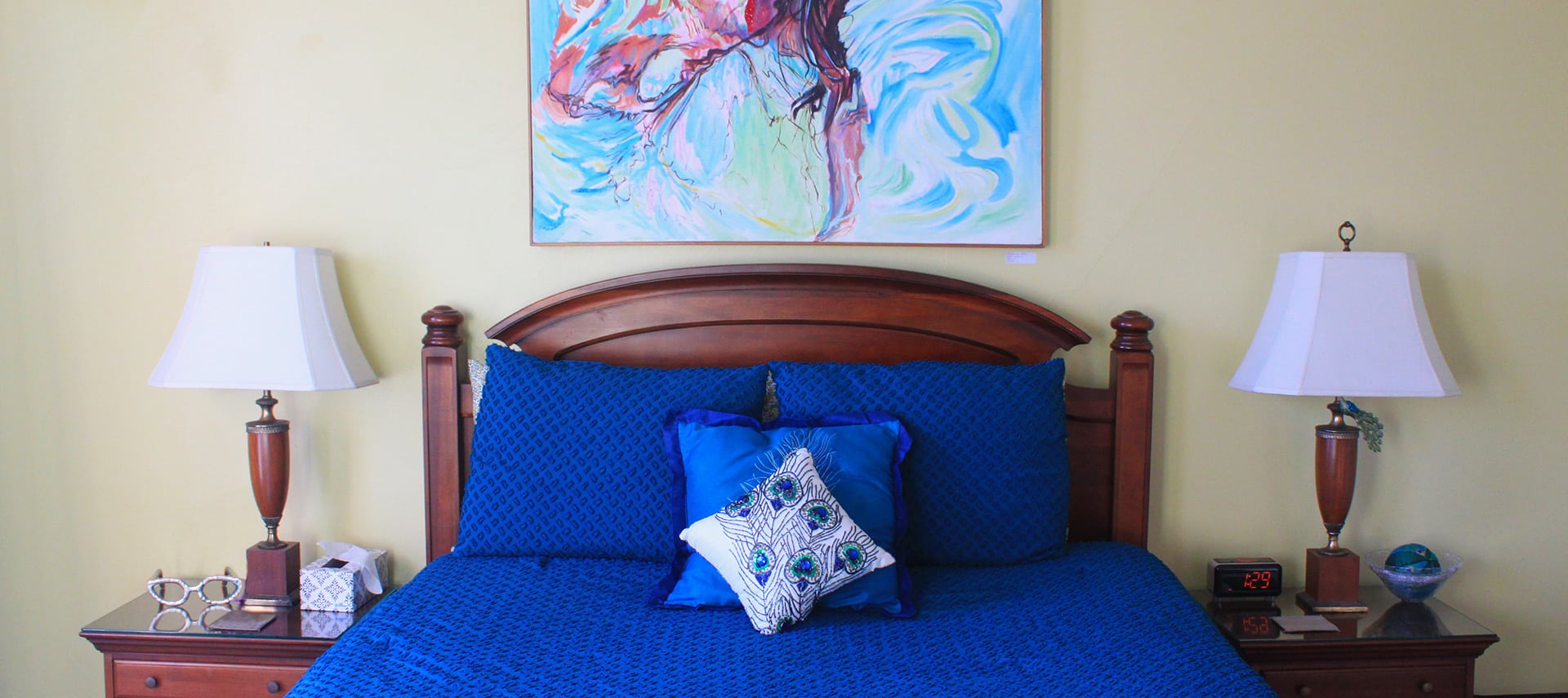A queen bed with dark brown headboard and peacock blue bedspread in a room with pale yellow walls. A painting of a woman in a pool hangs on the wall above the headboard.
