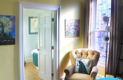 A yellow upholstered chair with a pillow printed with a peacock in a room with yellow walls, white woodwork, a stained glass window, a door leading into a light green bathroom. To the right of the chair is a lamp and a book shelf displaying books and art objects, and there are paintings on the walls.