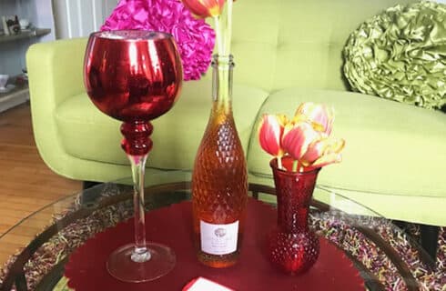 Rose wine bottle, tulips in a vase, and a large decorative wineglass-shaped candleholder on a red placemat on a coffee table in front of a light green couch with a fluffy pillow.