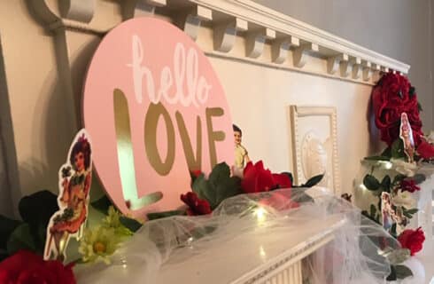 An ornate white carved wood fireplace mantel decorated with romantic decorations including red roses, white tulle, a sign that says Hello Love, and small engraved cutout Victorian pictures of people.
