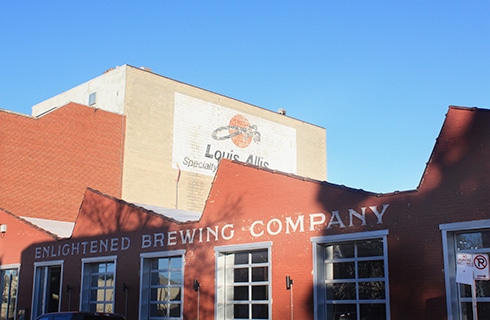 Brick facade of Enlightened Brewing Company with angular roofline, large metal-framed windows. Louis Allis warehouse behind the brewing company.