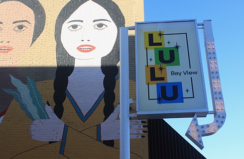 Brick building painted with a mural of women, with a sign that says LULU Bay View and has an arrow with lightbulbs.