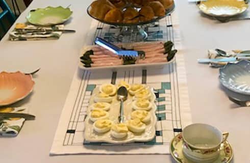 dining table set for breakfast with colorful and whimsical plates and silverware, a linen tablecloth. Deviled eggs and meat on platters, and a serving stand with baked goods.