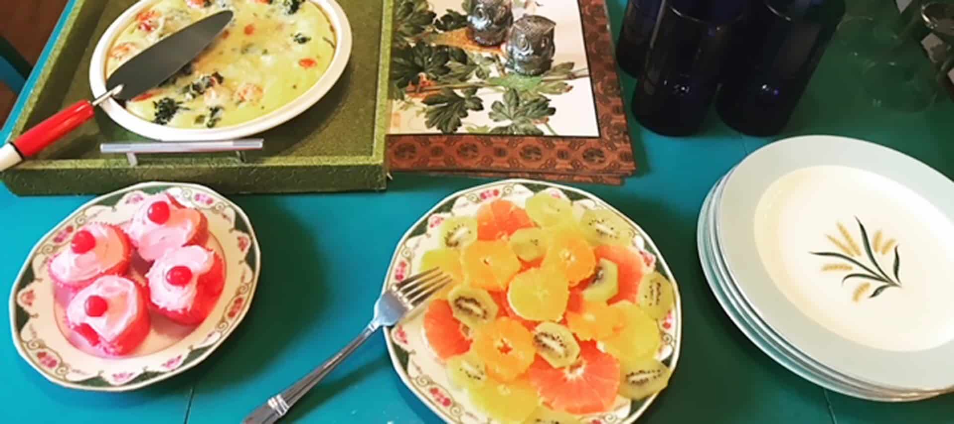 Breakfast tray and plates with quiche, fruit salad, and iced cookies, with china plates and glasses.