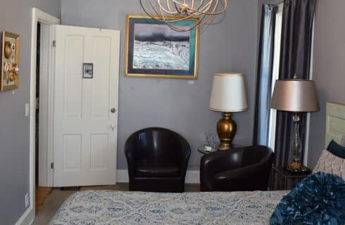 Bedroom with grey walls, dark brown upholstered chairs, two lamps, and a queen bed with a patterned bedspread, a headboard made from ajn antique door, and a deocrative pillow.