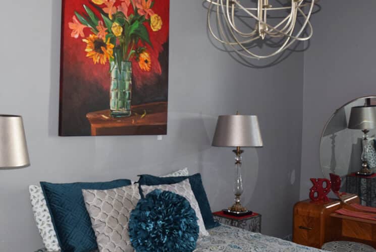 Bedroom with grey walls with a red painting of a vase of flowers, a bed with a patterned bedspread and blue and white pillows, a silver lamp on a nightstand, and a metal chandelier.