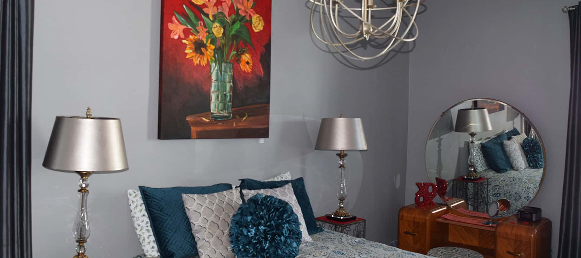 Bedroom with grey walls with a red painting of a vase of flowers, a bed with a patterned bedspread and blue and white pillows, a silver lamp on a nightstand, and a metal chandelier.