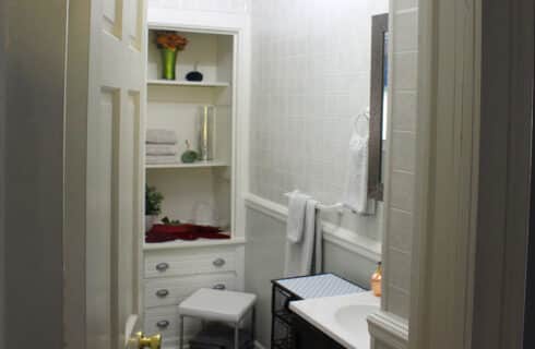 A bathroom with built in cabinet with open shelving and drawers, a stool, a towel bar, sink and mirror