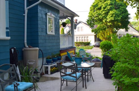 Patio in the side yard of the blue Victorian house, with several tables and chairs, plants, bushes, flowers and trees.