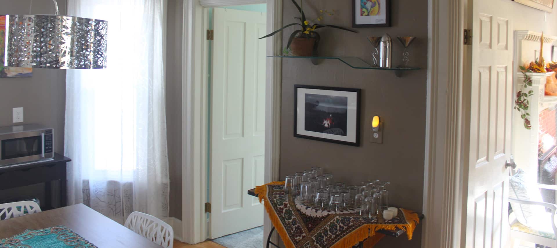 Kitchenette and dining area with a microwave, table, chairs, cart with glassware, a decorative silver metal ceiling lamp, dark beige walls with a variety of artwork, and white woodwork and doors into other rooms.