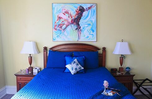 Guestroom with beige walls with a queen bed with dark brown headboard, blue bedspread, and two lamps on nightstands. A painting on the wall above the headboard depicts a woman floating in a swimming pool.