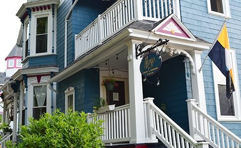 Large blue Victorian house with shutters, porch with white railings leads to the front door. The Muse Gallery Guesthouse sign and Milwaukee flag hang from the columns of the front porch.