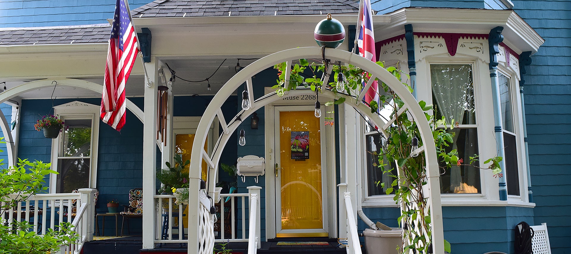 Yellow entrance door with the words Muse 2268 on the white trim above it. Blue siding on large Victorian house. A white arched arbor with vines frames the porch steps and railings leading to the yellow door.