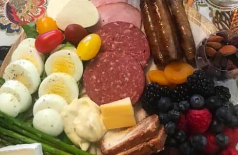 A tray of sliced hard boiled eggs, various meats and cheese and fruit on a tabletop with a patterned tablecloth.