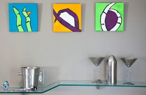 Bright abstract art pieces over a funky glass shelf holding silver cocktail items.