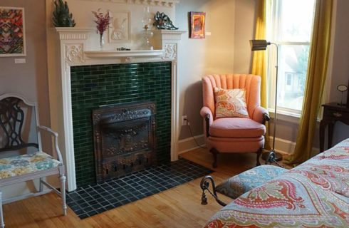 Tiled fireplace is the focal point of a bedroom with a bed, armchairs and a vanity.