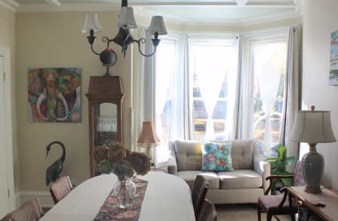 Dining room with tan walls, a dining table with a beige table cloth, fabric runner and vase of dried hydrangeas. Artwork on the walls, a chandelier, large bay window with white curtains. A dark brown china cabinet and a couch. A sideboard wtih a lamp and objects.