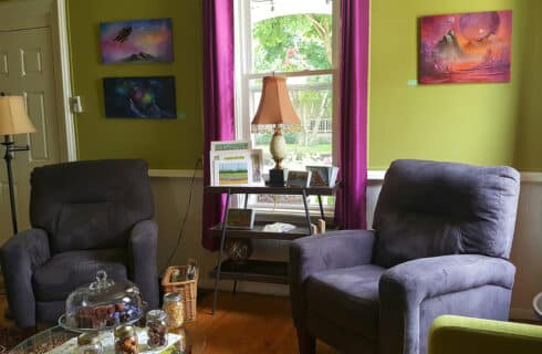 Lounge with bright green walls and purple curtains on a large window, colorful artwork on the walls. Dark purple cushioned armchairs. A side table with lamp. A coffee table with snacks.