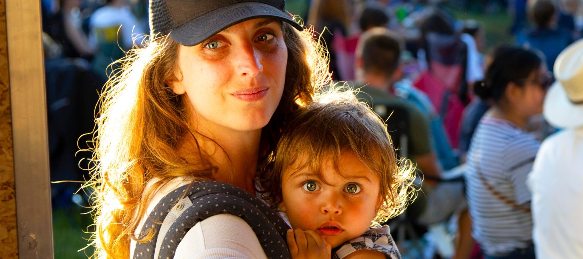 Woman with black ball cap and long hair holding a young girl in front of a crowd