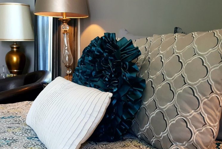 Bed made up with tan and teal paisley spread and decorative pillows next to a nightstand and leather club chair.