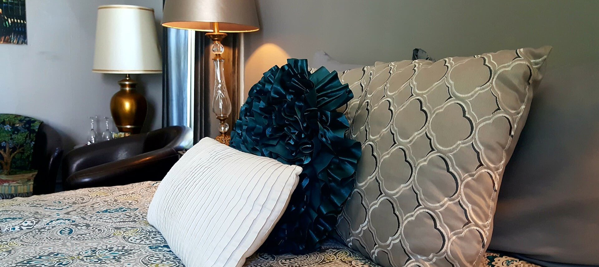 Bed made up with tan and teal paisley spread and decorative pillows next to a nightstand and leather club chair.