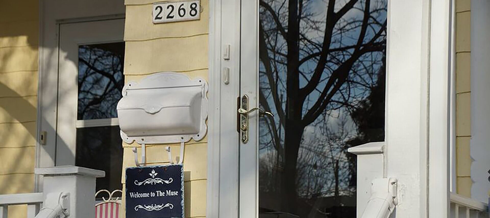 Front door of yellow house with white metal mailbox and sign that says, Welcome to The Muse.
