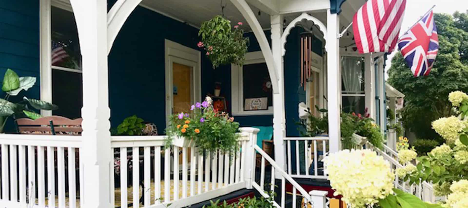 White wooden side porch with vertical stair railings and railings. Arched decorative wooden trim on the porch columns. Blue sliding, white window and door trim, yellow door, flower planters on the porch railing and hanging flower baskets.