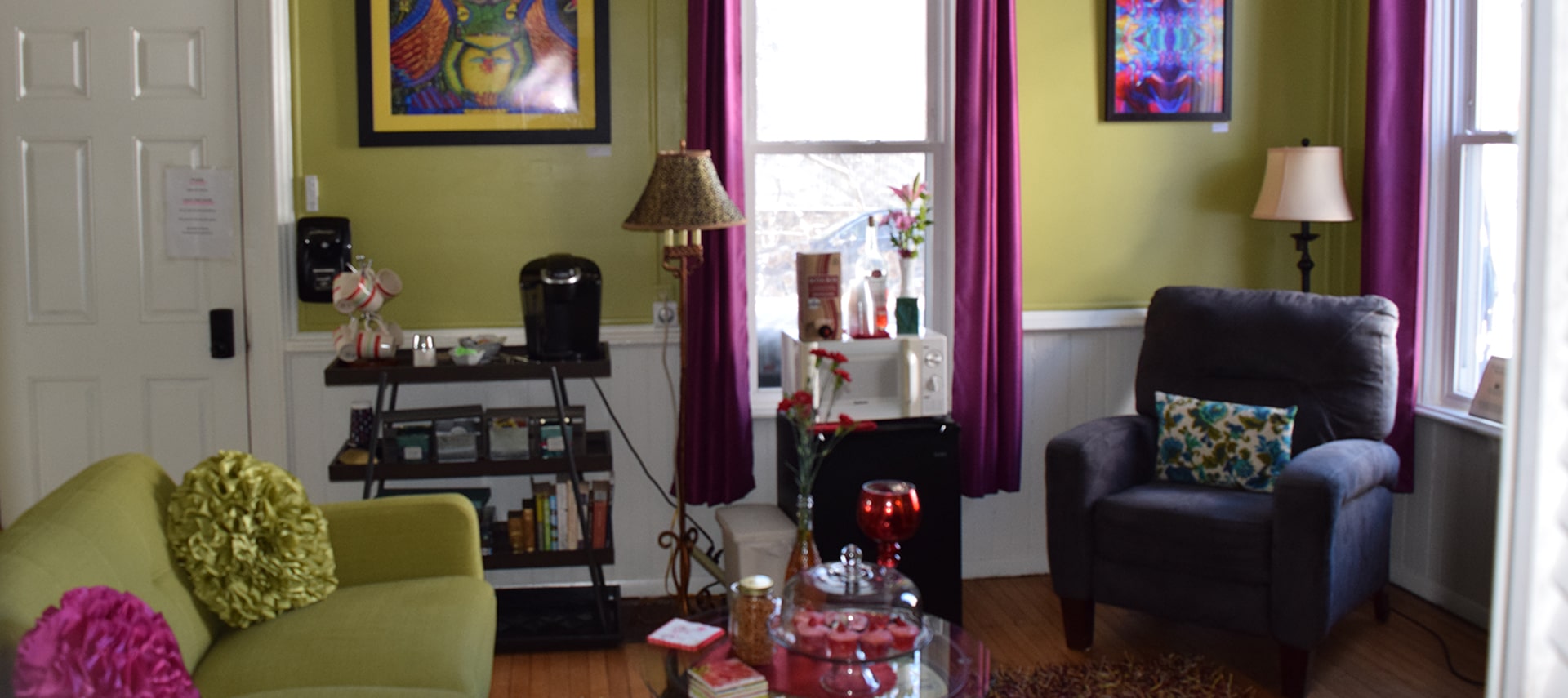 Lounge area with a couch and upholstered chair, coffee table and coffee maker, microwave. Artwork on green walls, white wood trim and door, and a large window with purple curtains.