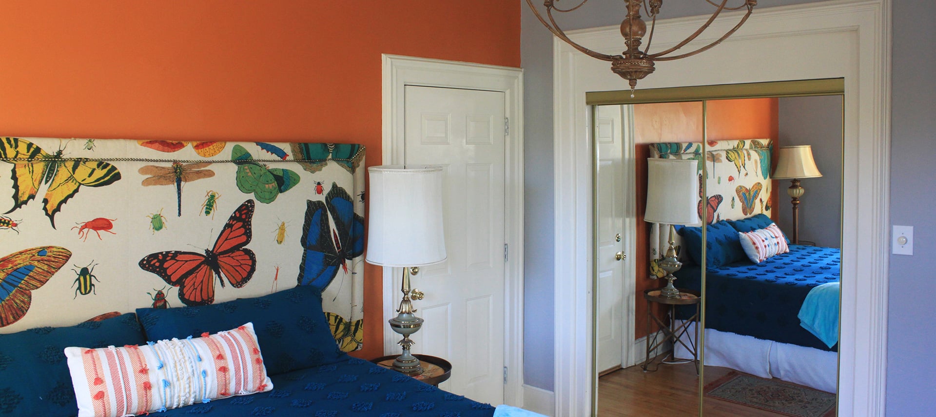A unique bedroom with orange walls, white trim, an artistic headboard with a butterfly design, a blue bedspread and a decorative pillow.