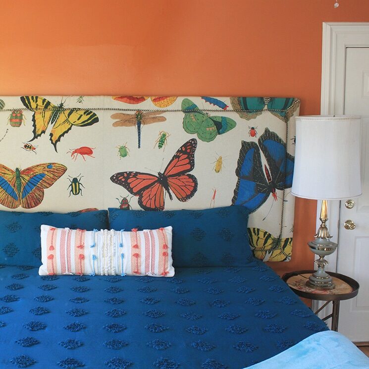 Guestroom with orange walls with a queen bed with a blue bedspread and a nightstand with a lamp.