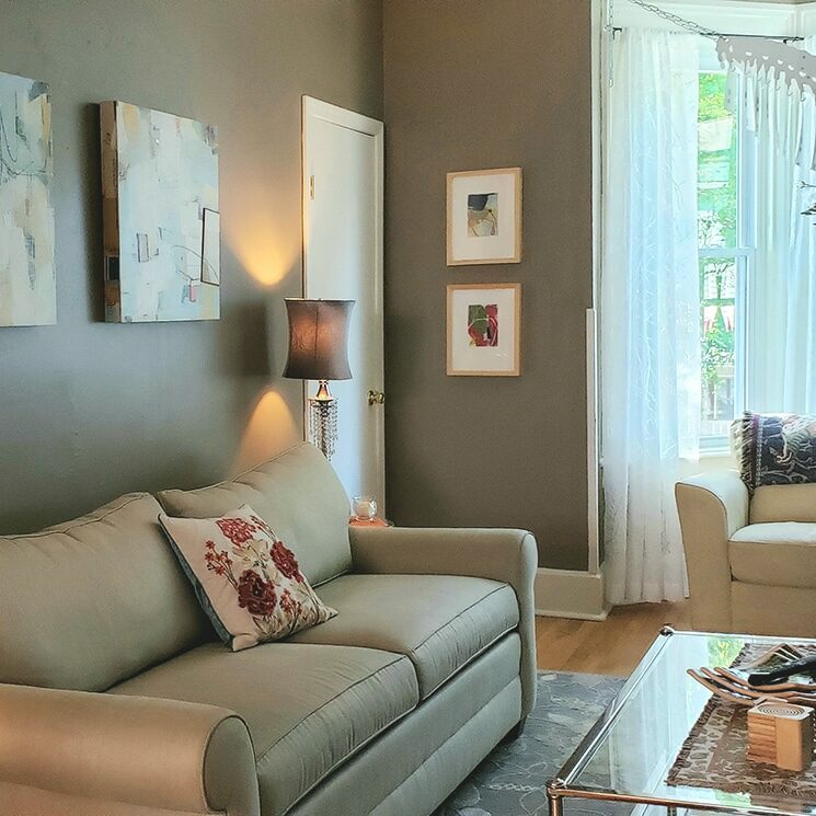 Beige sofa, with glass and chrome coffee table in a lounge area with grey walls, artwork and sheer white curtains on a large window.