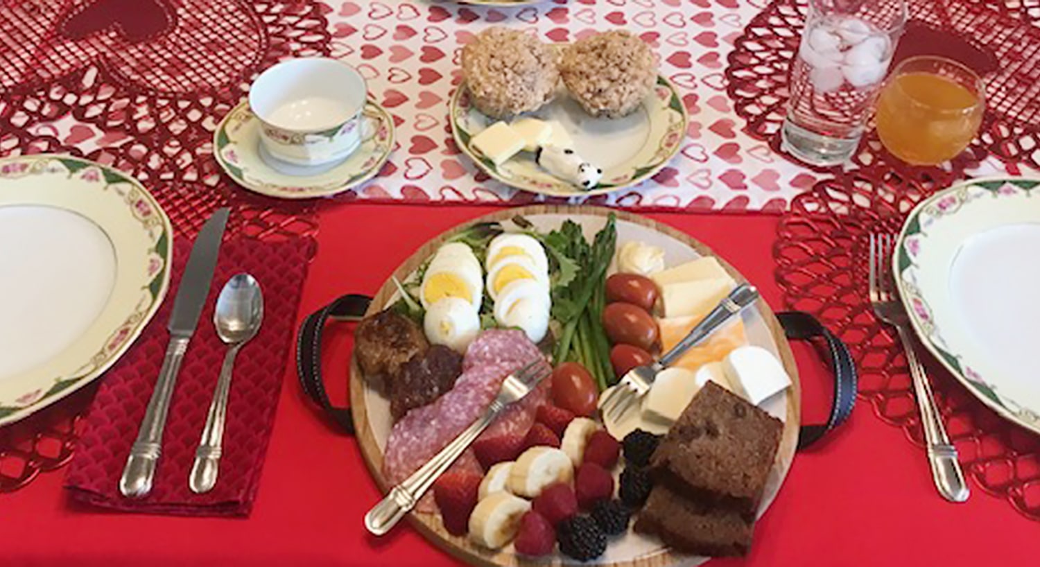 Place setting for a Valentine's meal - a plate full of meat, cheese, eggs, fruit, vegetables, and bread, a dessert plate with baked goods, and a teacup on a table with a red tablecloth.