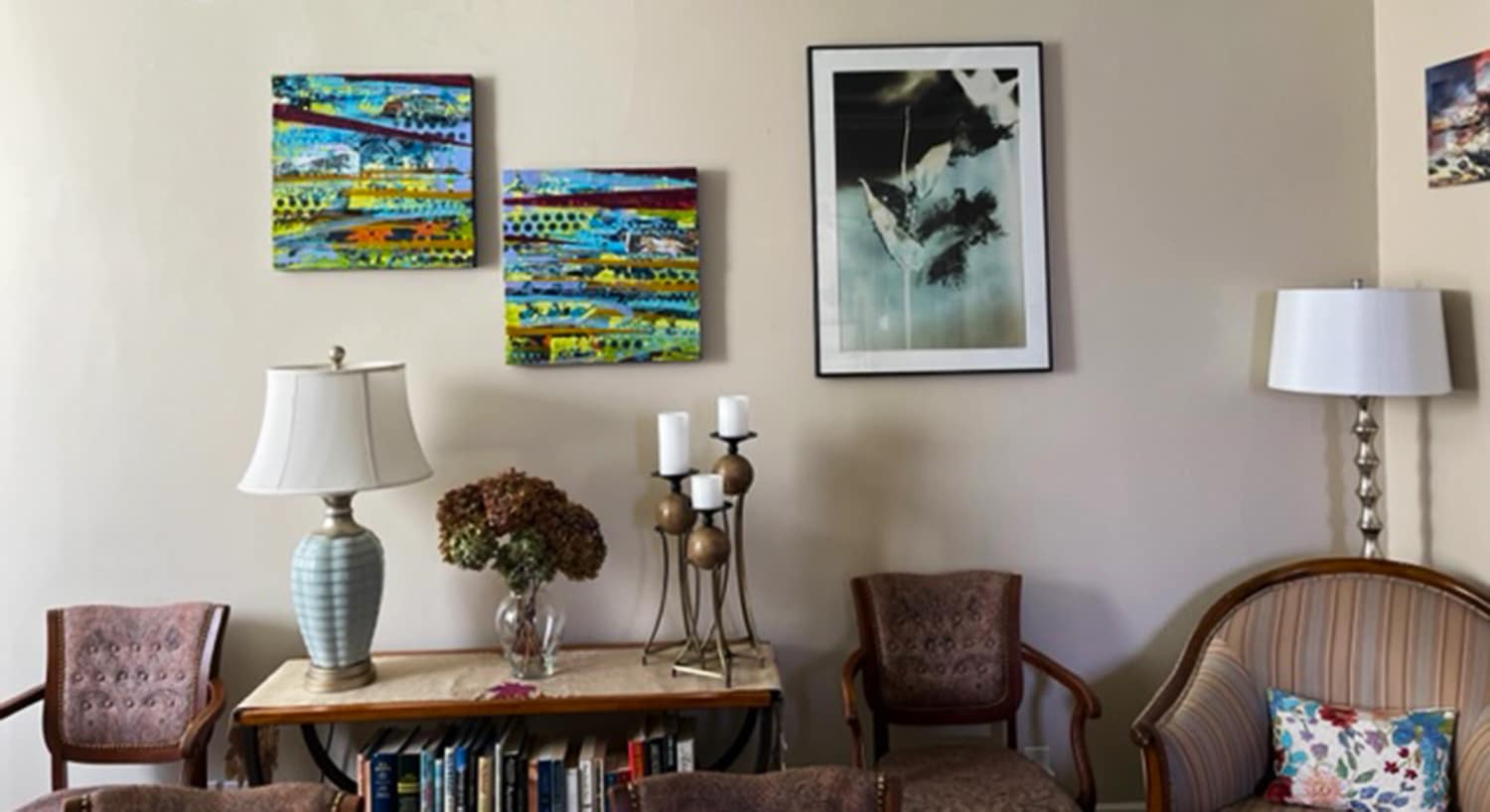 Colorful abstract paintings hang on the light tan walls. A sideboard holds a lamp and candleholder. Two armchairs and an upholstered chair.