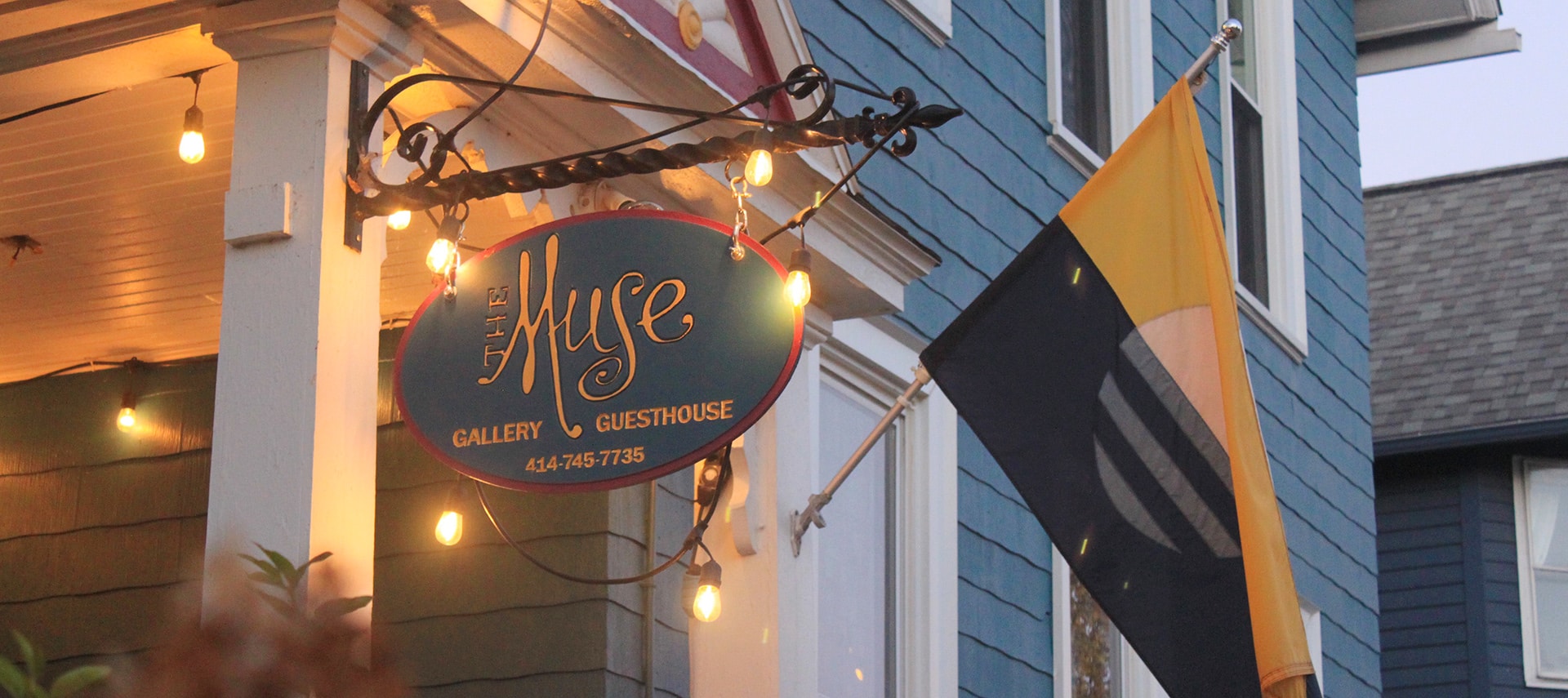 Detail of exterior front entrance with white string lights on white porch columns, a hanging carved sign that reads The Muse Gallery Guesthouse 414-745-7735 and the Milwaukee flag. Blue siding and white trim on the house.