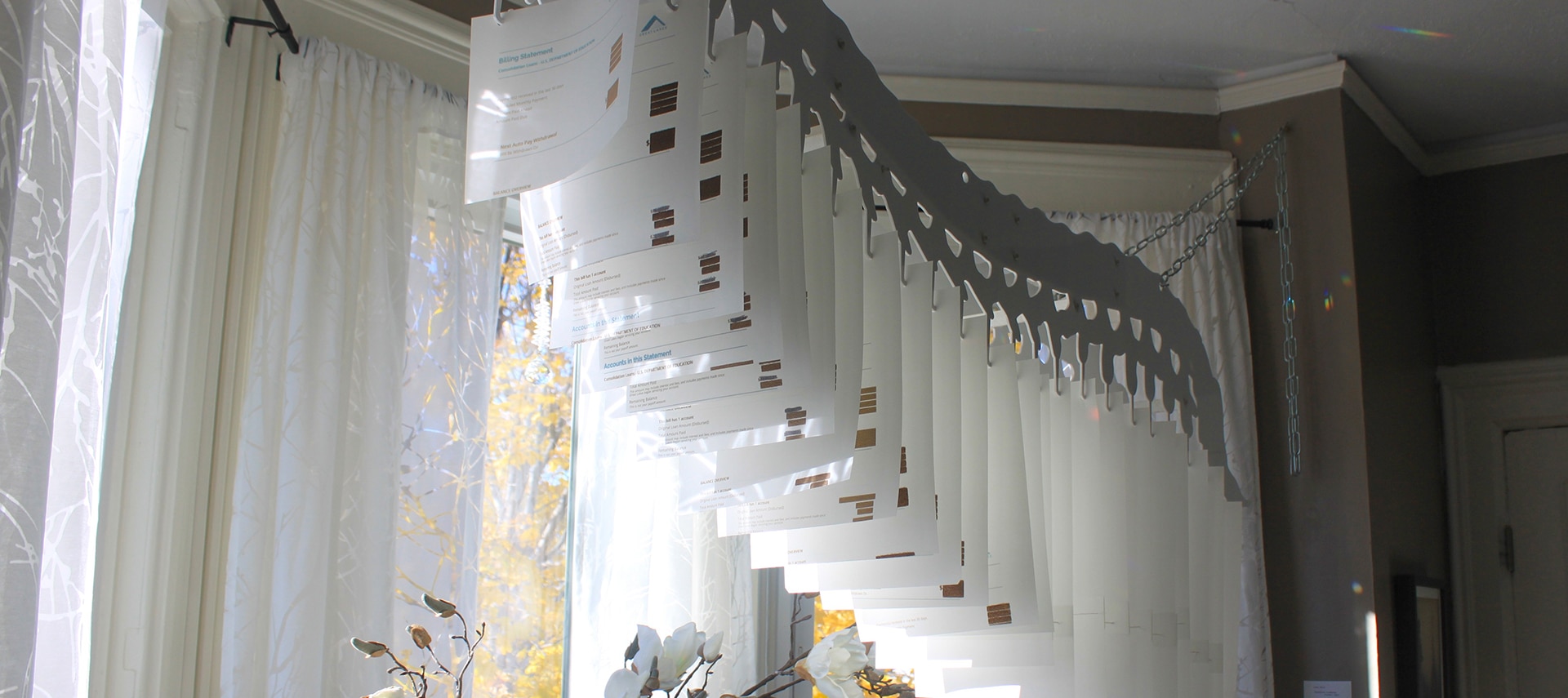 A hanging paper sculpture in a bay window of a room with tan walls. Flowers in a vase below the sculpture.