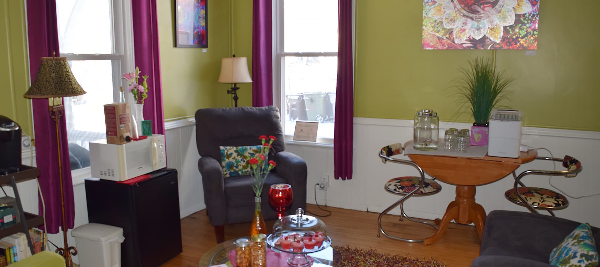 Lounge with bright green walls, large windows with purple curtains, eclectic furniture including an upholstered purple chair, lamps, a coffee table, and a small table with two chairs.