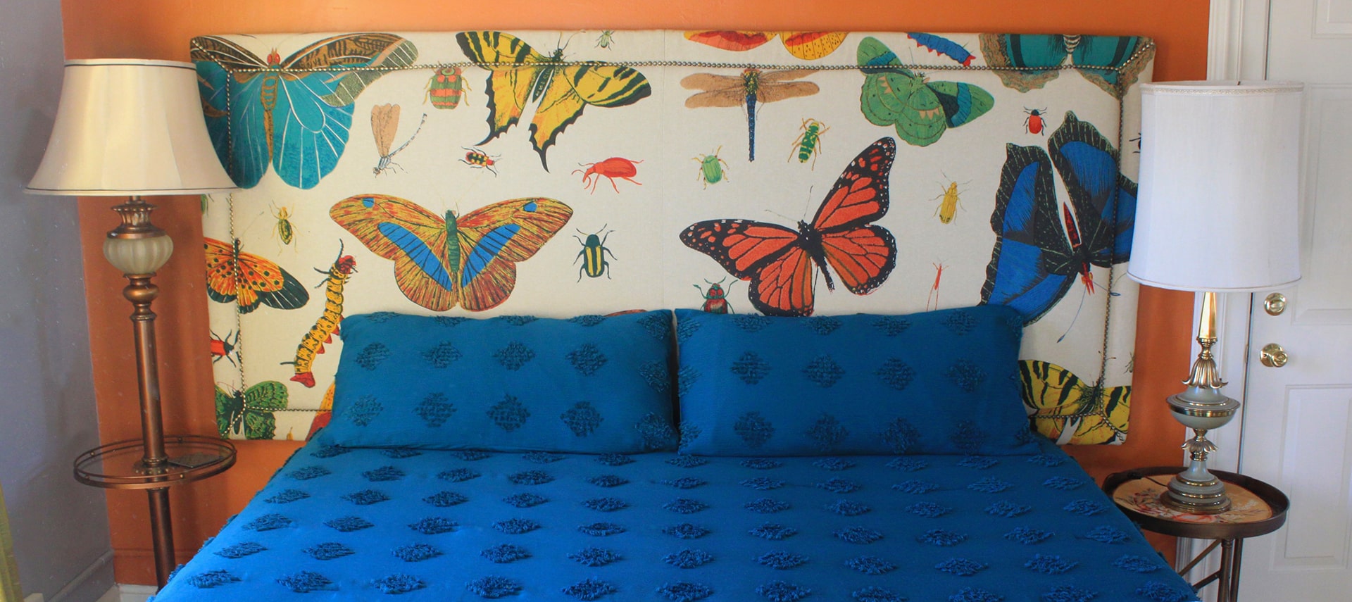 A queen-sized bed with blue bedspread and pillows, a headboard with butterfly pattern, and 2 lamps, in a room with orange walls.