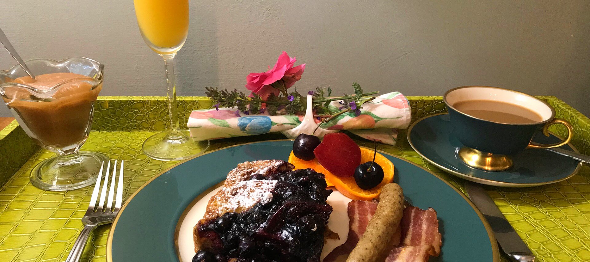 French toast with blueberry compote accompanied by sausage and bacon on a teal and white china plate. 