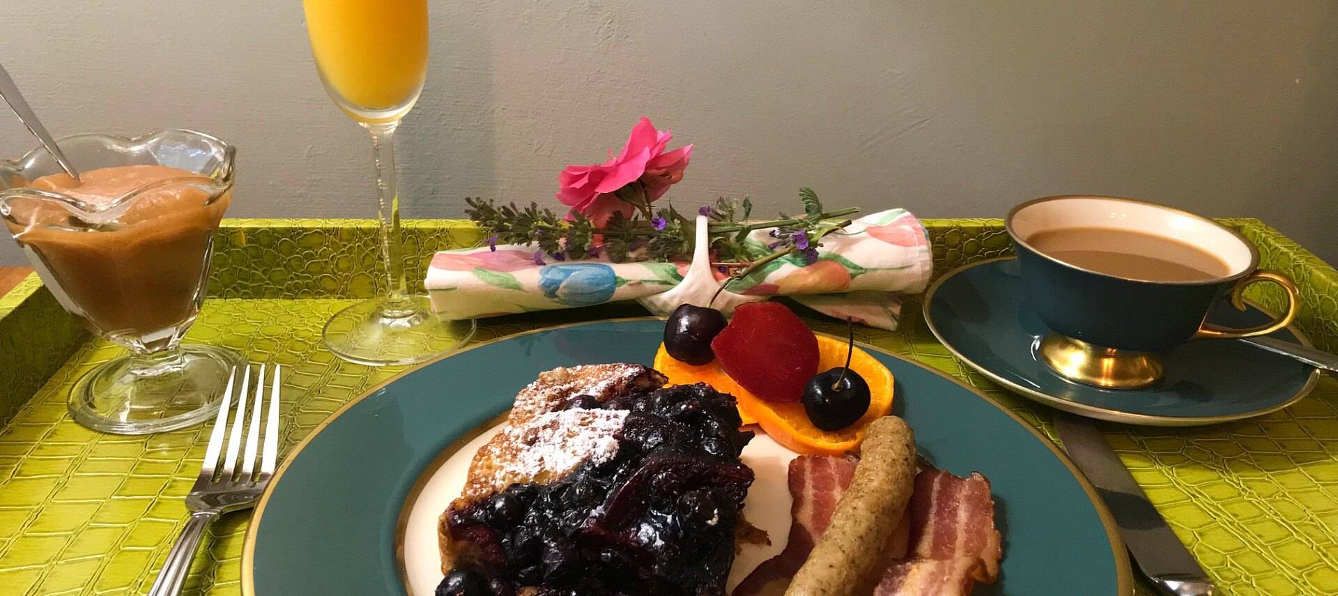 French toast with blueberry compote accompanied by sausage and bacon on a teal and white china plate. 